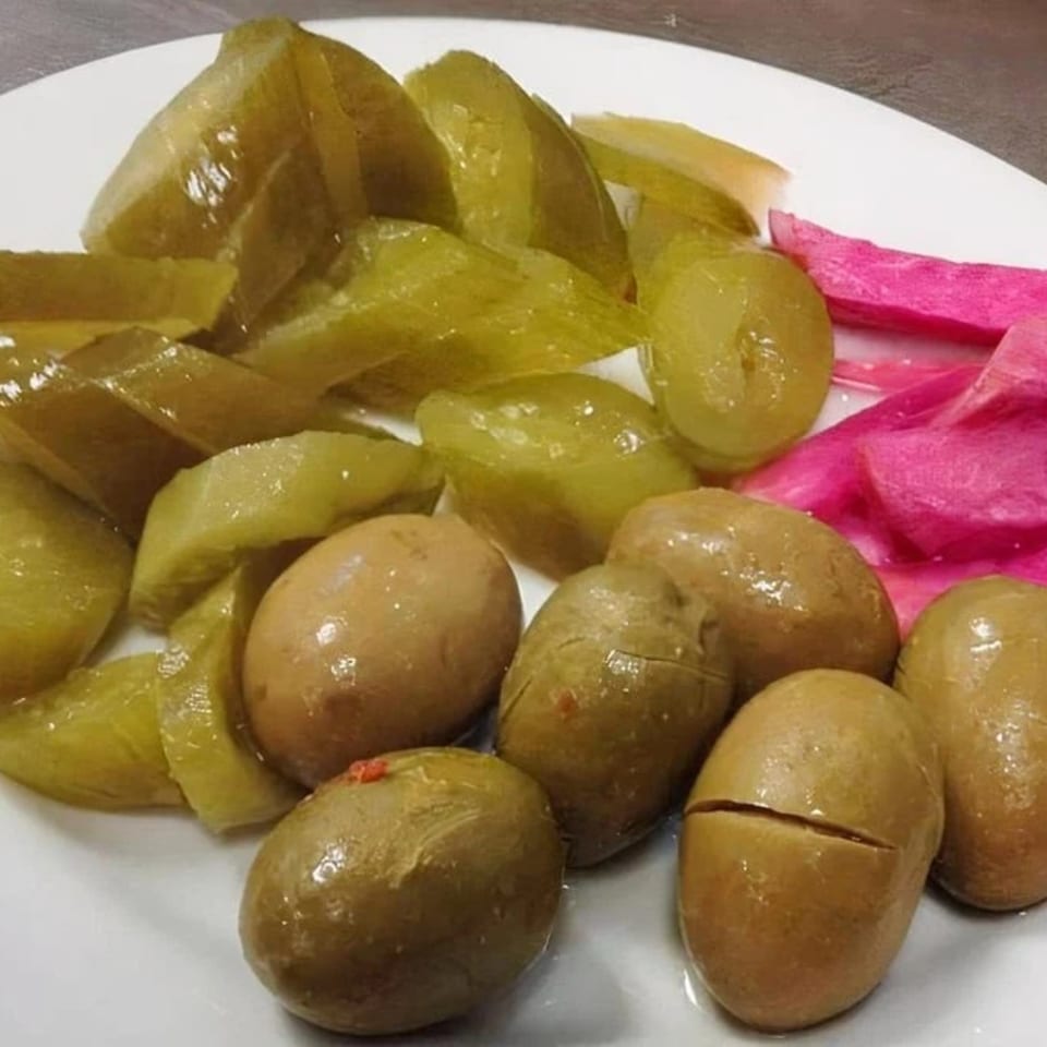 A white plate holds green pickles, whole olives, and bright pink pickled turnips.